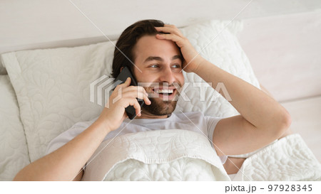 Cheerful shocked young caucasian guy lies on bed, wakes up, calls by phone, looks at free space in bedroom 97928345