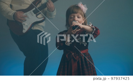 Little girl on stage in vintage dress, she sings into microphone and dances, her father plays an electric guitar. Color music is shining and smoke is billowing. Family time, creativity and hobbies Little girl on stage in vintage dress, she sings into microphone and dances, her father plays an electric guitar. Color music is shining and smoke is billowing. Family time, creativity and hobbies 97937425