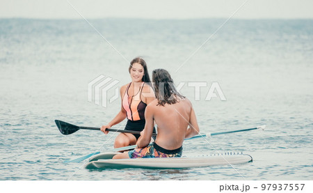Sea woman and man on sup. Silhouette of happy young woman and man, surfing on SUP board, confident paddling through water surface. Idyllic sunset. Active lifestyle at sea or river. 97937557