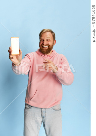 Smilling bearded man in casual style clothes showing empty smartphone screen over blue background. Concept of emotions, happiness, ad, sales. 97941659