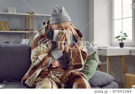 Old man is sitting at home, warmly wrapped in warm blanket, dressed in knitted sweater and hat. 97943384