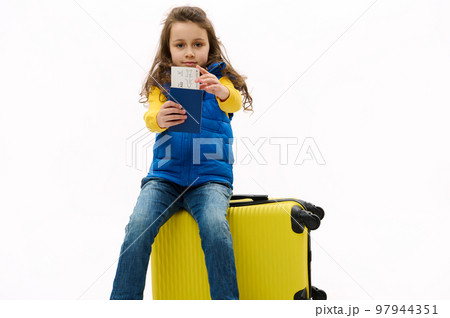 Isolated portrait on white background of a Caucasian charming kid girl in blue jacket, holding out an air ticket and boarding pass at camera, sitting on yellow travel bag, going for weekend getaway. 97944351