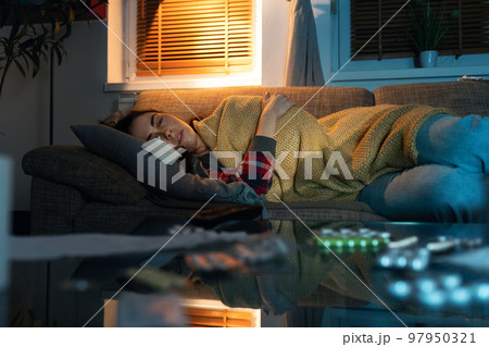 Depressed young Caucasian woman covered blanket lies on sofa and watching tv. Close up view from table with pills. Concept of psychology disorder 97950321