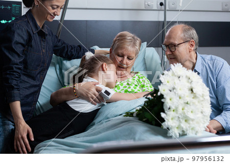 Little granddaughter giving flowers bouquet to sick grandmother. Old female patient receiving floral present from relatives on hospital visit. Girl hugging sick old woman at medical clinic bedroom. Little granddaughter giving flowers bouquet to sick grandmother. Old female patient receiving floral present from relatives on hospital visit. Girl hugging sick old woman at medical clinic bedroom. 97956132