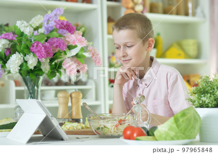 little boy eating salad and looking at tablet screen on kitchen table at home 97965848