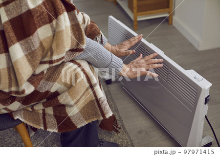 Senior man at home warms his hands on electric heater due to problems with central heating. 97971415