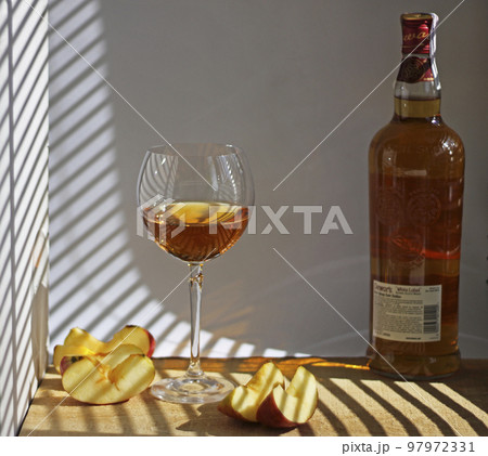 Still life with a glass of wine, apple slices and a bottle. There is a shadow from horizontal blinds on the wooden table and walls. Modern still life with apples. Still life with a glass of wine, apple slices and a bottle. There is a shadow from horizontal blinds on the wooden table and walls. Modern still life with apples. 97972331