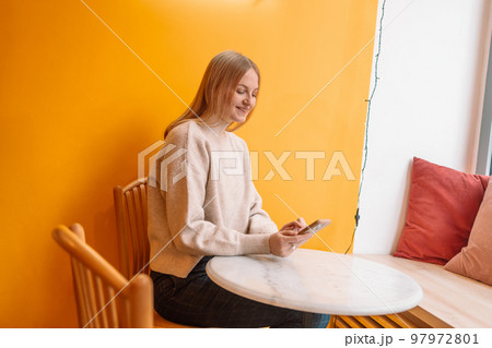 Attractive young blonde smiling woman with cup of coffee using mobile phone in cafe on a yellow background 97972801