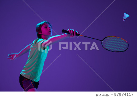 Portrait of teen boy in uniform playing badminton, hitting shuttlecock with racket isolated over blue purple background in neon ligth Portrait of teen boy in uniform playing badminton, hitting shuttlecock with racket isolated over blue purple background in neon ligth 97974117