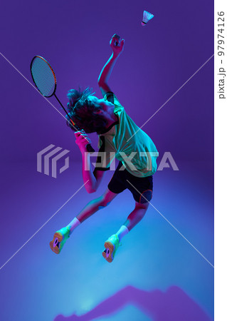 Dynamic game. Teen boy in uniform playing badminton, hitting shuttlecock in a jump over blue purple background in neon ligth 97974126