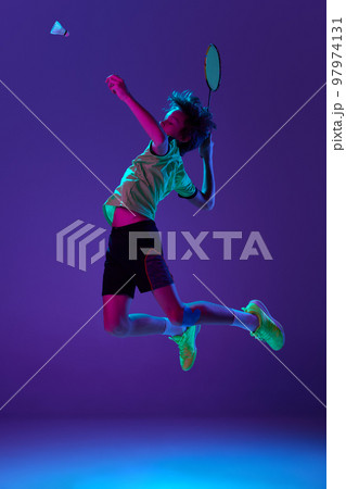 Concentrated teen boy in uniform playing badminton, hitting shuttlecock in a jump over blue purple background in neon ligth 97974131