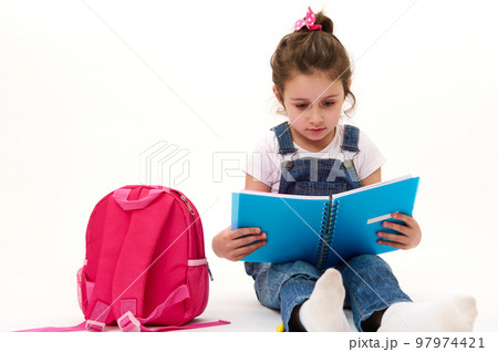 Caucasian lovely child, little girl in blue denim overalls and t-shirt, first grade student reading textbook, sitting near her pink backpack on white background. Copy ad space. Back to school concept 97974421