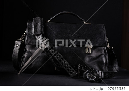 A large black knife and a watch on the background of a leather bag. Male style. Black items on black. A large black knife and a watch on the background of a leather bag. Male style. Black items on black. 97975585
