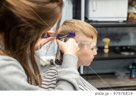 A schoolboy is sitting in a barbershop, doing his hair with a clipper A schoolboy is sitting in a barbershop, doing his hair with a clipper 97976819