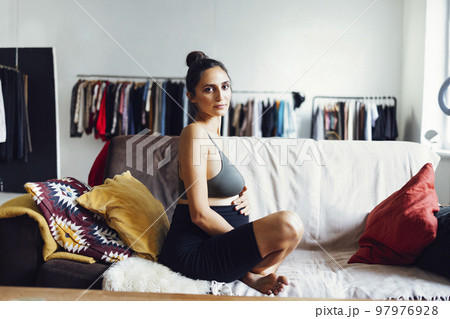 Cheerful young brunette latin pregnant sportswoman sitting on couch in light room 97976928
