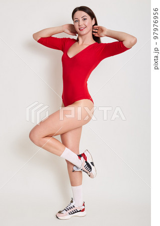 Close up photo of young fitness woman, being photographed for her social network, posing isolated over white background, female standing on one leg and wearing red stylish combidress. Sport concept. 97976956