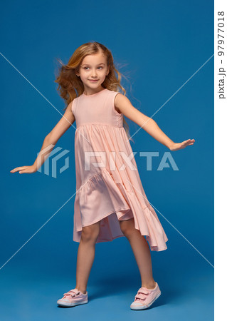 Fashionable little girl in a pink dress is posing on a blue background. 97977018