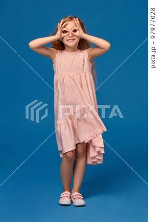 Fashionable little girl in a pink dress is posing on a blue background. 97977028