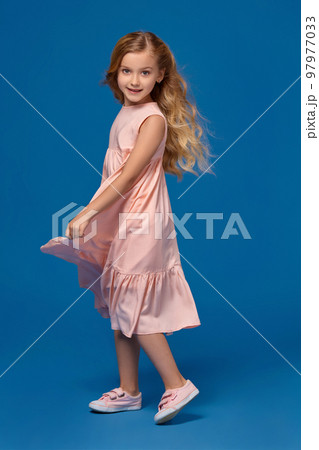 Fashionable little girl in a pink dress is posing on a blue background. 97977033