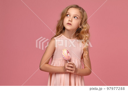 Little girl with a blond curly hair, in a pink dress is posing with a candy 97977288