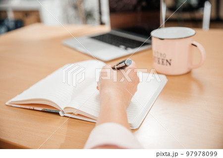 Close up of a woman's hands writing in a notepad placed on a wooden table, laptop, home office and work concept, plans and business meeting 97978099