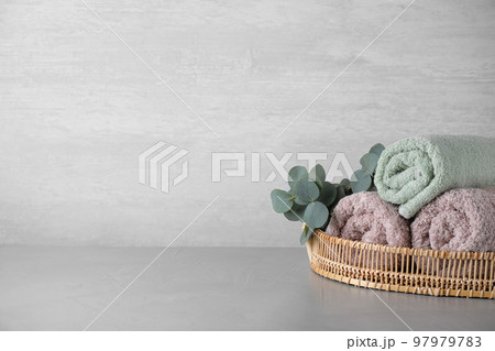 Wicker tray with rolled bath towels and eucalyptus branch on grey table. Space for text Wicker tray with rolled bath towels and eucalyptus branch on grey table. Space for text 97979783