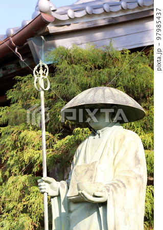 お寺の庭に立つ笠をかぶる親鸞聖人の銅像。背景にお寺と緑が映り込んだ晴れた日の写真。 97985437