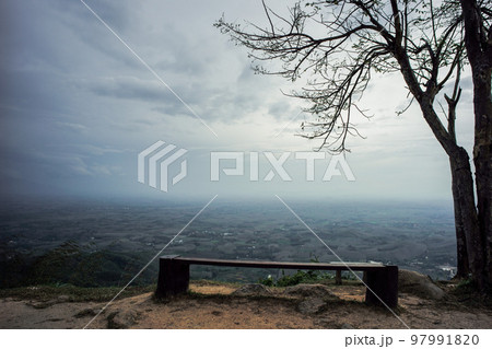 Empty wooden bench under the tree on the mountain with landscape and cloudy sky in background Empty wooden bench under the tree on the mountain with landscape and cloudy sky in background 97991820