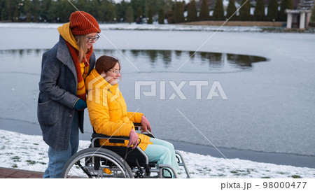Caucasian woman pushes her friend on a wheelchair by the lake. 98000477