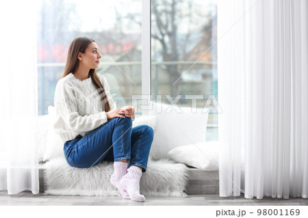 Young woman in sweater sitting near window indoors 98001169