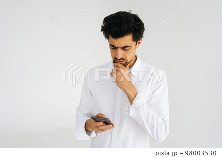 Studio portrait of thoughtful bearded business man in shirt holding smartphone, using online mobile apps, looking on device screen, chatting in messengers standing on white isolated background. Studio portrait of thoughtful bearded business man in shirt holding smartphone, using online mobile apps, looking on device screen, chatting in messengers standing on white isolated background. 98003530
