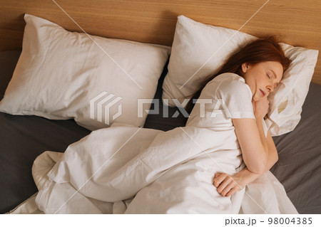 Top view of cute calm redhead young woman wearing soft comfortable night clothes sleeping in comfortable bed lying down on soft pillow. Peaceful pretty lady resting covered with blanket. Top view of cute calm redhead young woman wearing soft comfortable night clothes sleeping in comfortable bed lying down on soft pillow. Peaceful pretty lady resting covered with blanket. 98004385