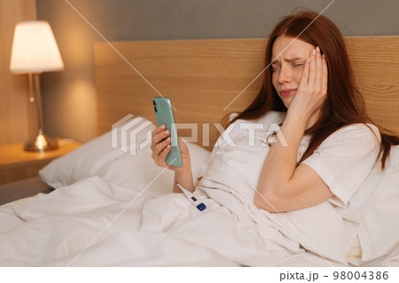 Crying ill red-haired young woman having online video call via smartphone lying in bed at home. Sick redhead female using cellphone to talk with family. Concept of fever, flue or virus. 98004386