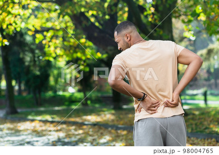 Tired man after jogging in park, hispanic man has severe pain in back and muscles after fitness, massaging his side with hand near trees. 98004765