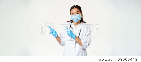 Excited asian female doctor points left, wears medical mask as covid-19 preventive measures, standing in uniform over white background Excited asian female doctor points left, wears medical mask as covid-19 preventive measures, standing in uniform over white background 98006448