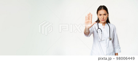 Asian doctor shows palm, stop gesture. Serious displeased female physician extend her arm, prohibit action, taboo sign, standing in uniform against white background Asian doctor shows palm, stop gesture. Serious displeased female physician extend her arm, prohibit action, taboo sign, standing in uniform against white background 98006449