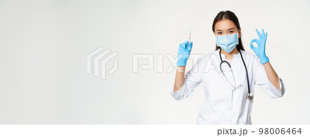 Covid-19 vaccination and healthcare. Asian woman doctor in medical face mask and gloves, holding vaccine syringe and show okay sign, white background 98006464