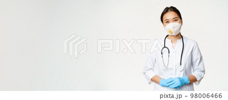 Smiling asian female doctor in respirator, medical mask and rubber gloves, ready to help, standing in clinic uniform over white background. Covid-19 and quarantine concept 98006466
