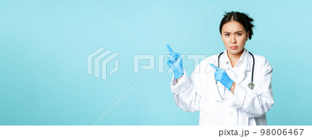Angry female doctor or nurse, pointing fingers at upper left corner with disapproval, sulking and furrow eyebrows, standing in medical uniform, blue background Angry female doctor or nurse, pointing fingers at upper left corner with disapproval, sulking and furrow eyebrows, standing in medical uniform, blue background 98006467