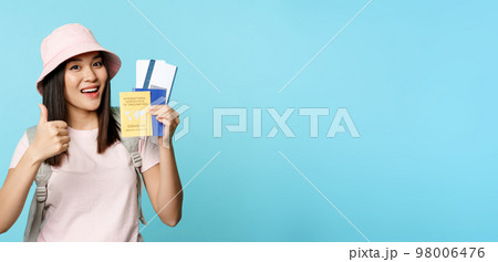 Smiling happy asian tourist, girl shows thumbs up and international vaccination certificate, vaccine passport and tickets, standing over blue background 98006476