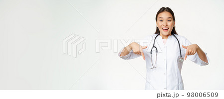 Enthusiastic medical staff, asian female healthcare worker, pointing fingers down and smiling amazed, showing discounts, sale in clinic, white background 98006509