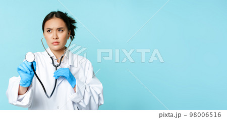 Image of serious medical worker, asian doctor listening to patient lungs with stethoscope, examining person, standing over blue background 98006516