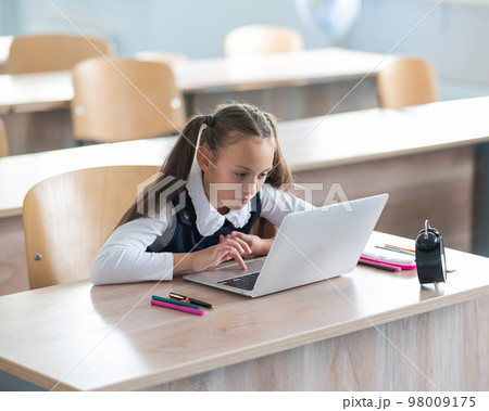Caucasian girl sits at a desk at school and studies at a laptop. Alarm clock on the table. 98009175