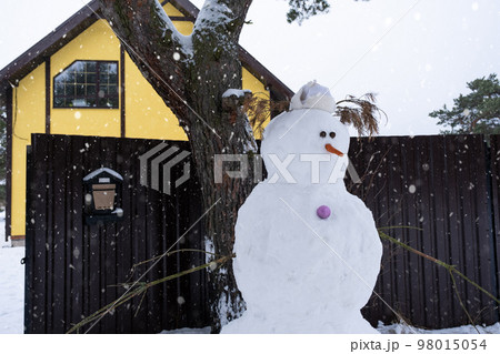 Funny snowman in a hat and scarf on the background of a yellow house in the yard. Winter, winter entertainment, snowfall 98015054