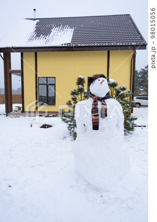 Funny snowman in a hat and scarf on the background of a yellow house in the yard. Winter, winter entertainment, snowfall 98015056
