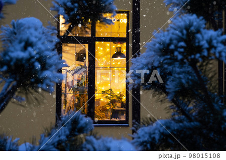 Cozy festive window of the house outside with the warm light of fairy lights garlands inside - celebrate Christmas and New Year in a warm home. Christmas tree, bokeh, snow on pine trees and snowfall 98015108