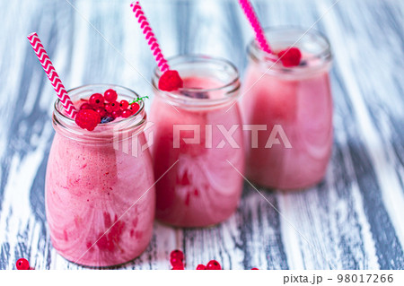 Front view of three berries milkshakes in glasses jars with ingredients on wooden table Front view of three berries milkshakes in glasses jars with ingredients on wooden table 98017266