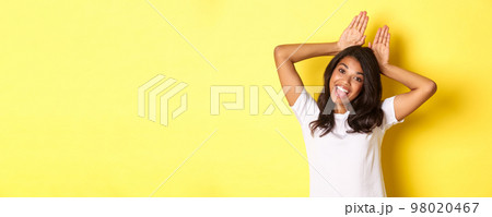 Portrait of attractive african-american girl, smiling and showing bunny ears on head, standing silly and coquettish over yellow background 98020467