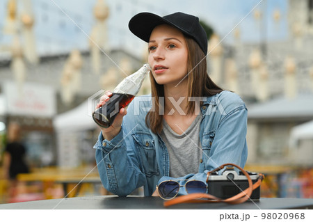 Caucasian female traveller drinking cola from glass bottle while sitting at outdoor street food restaurant 98020968