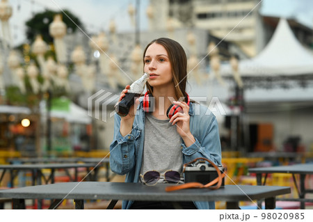 Caucasian female traveller drinking cola from glass bottle while sitting at outdoor street food restaurant 98020985